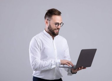 Ofisteki olumlu genç adam gri stüdyo üzerinden dizüstü bilgisayar kullanıyor.