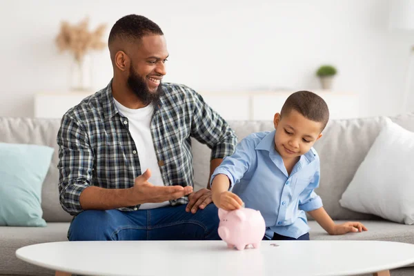 Schwarz junge und seine papa putting geld im sparschwein drinnen — Stockfoto
