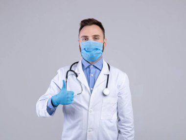 Laboratuar önlüklü, koruyucu maskeli ve lastik eldivenli bir doktor gri stüdyo geçmişinde baş parmak hareketlerini gösteriyor.