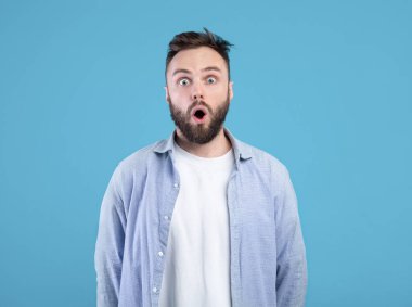 Afallamış genç bir adamın portresi mavi stüdyo arka planında şok içinde ağzını açıyor.