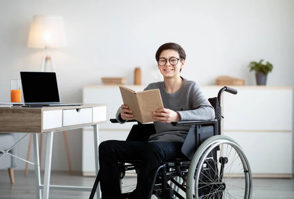 Happy disabled teenager in wheelchair reading book, doing online home assignment, space for design on laptop screen