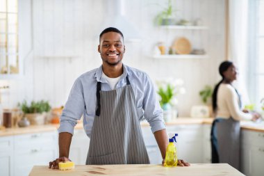 Önlüklü siyah bir adam portresi ve mutfakta kameraya gülümseyen lastik eldivenler, arka planda bulaşıkları yıkayan karısı.