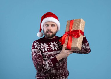Noel kıyafetli ve Noel Baba şapkalı genç adam mavi stüdyo arka planında hediye kutusunun içinde ne olduğunu dinliyor.