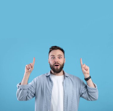 Yakışıklı sakallı adam şok olmuş yüz ifadesiyle mavi stüdyo arka planında boş bir yeri işaret ediyor.