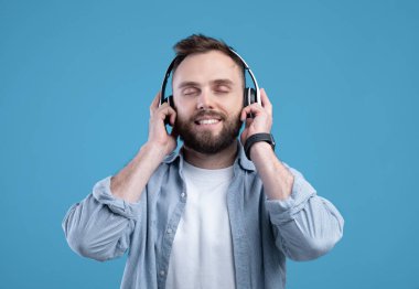 Sıradan giyinmiş, müzik dinleyen, kulaklık takan, mavi stüdyo geçmişli çekici sakallı bir adam.