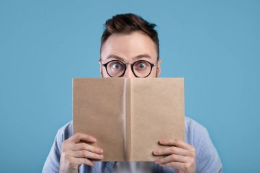 Gözlüklü genç adam şaşırdı kitap okuyor, gözlerini açıyor mavi stüdyo arka planda şaşkınlıkla
