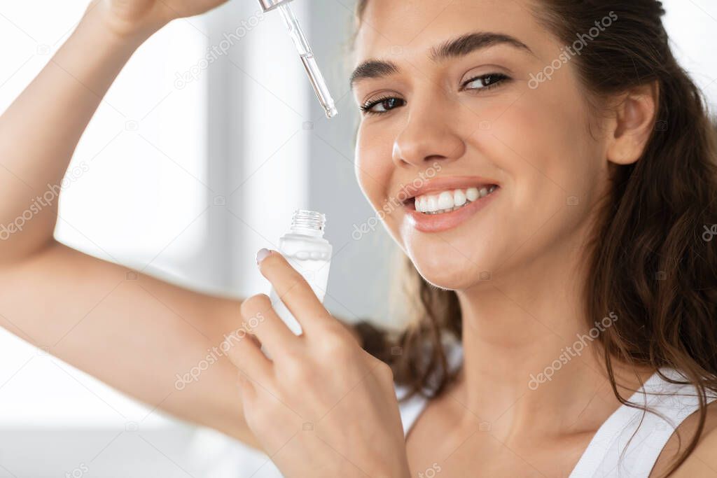 Happy Woman Applying Face Serum Posing Holding Dropper In Bathroom