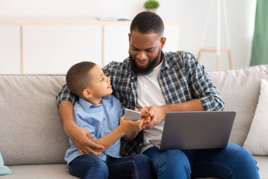 Siyah çocuk ve baba evde akıllı telefon ve dizüstü bilgisayar kullanıyor.