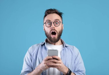 Şok olmuş sakallı adam mavi stüdyonun arka planında cep telefonuna bakıyor.