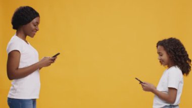Afrika kökenli Amerikalı anne ve kız akıllı telefonlara göz gezdiriyorlar, turuncu stüdyo arka planı ve aralarında boşluk var.