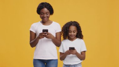 İnternet nesli. Afrikalı Amerikalı kadın ve kızı cep telefonlarıyla internette geziniyor, turuncu arka planda duruyorlar.