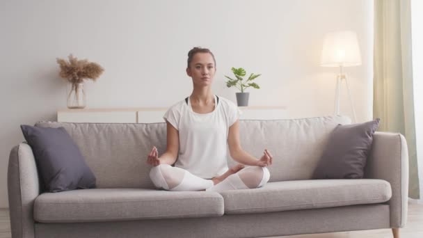 Молода усвідомлена жінка, що медитує вдома, сидить на дивані в положенні лотоса і дихає — стокове відео