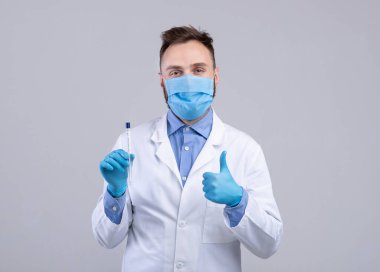 Maskeli ve eldivenli doktorun portresi kovid-19 PCR test tüpü taşıyor ve gri arka planda baş parmak hareketlerini gösteriyor.