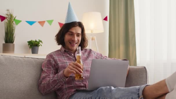 Młody szczęśliwy mężczyzna z butelką piwa wideo rozmowy z przyjaciółmi, taniec online, siedzi na kanapie w domu, zwolnione tempo — Wideo stockowe