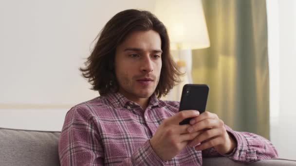 Evdeki kanepede oturan genç adama yakın çekim, akıllı telefondan sosyal medyada sörf yapmak. — Stok video
