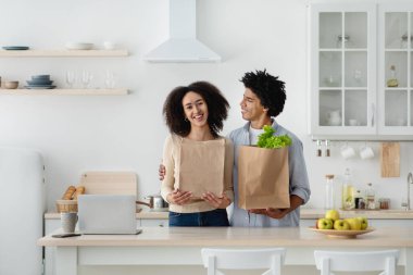 Mutlu, çekici, Afro-Amerikalı bir adam gülümseyen bir eşe bakıyor. Elinde kağıt torba ve sebzeyle.