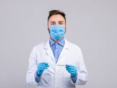 Tıbbi maske ve lastik eldiven takan genç bir doktor gri stüdyo arka planında koronavirüs PCR test tüpü tutuyor.