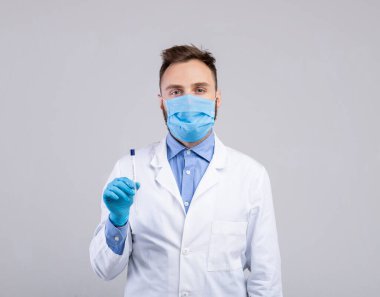 Korunaklı maske takan erkek doktor ve eldivenler, gri arka planda koronavirüs PCR testi için steril çubuk tutuyor.