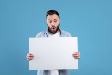 Mavi stüdyo arka planında beyaz afiş tutan şaşkın sakallı adam, reklam ya da tasarım için maket