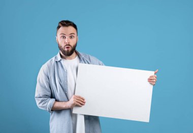 Şaşırdım genç adam boş beyaz poster gösteriyor, mavi stüdyo arkaplan, tasarım için alan sunuyor