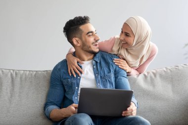 Başörtülü kadın yakışıklı kocasına dizüstü bilgisayarla sarılıyor.