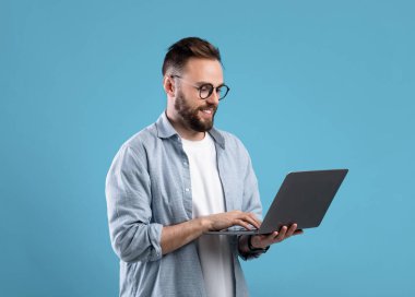 Gözlüklü kendine güvenen genç adam online iş için dizüstü bilgisayar kullanıyor ya da mavi stüdyo geçmişi ile iletişim kuruyor.