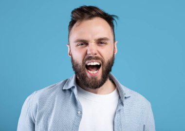 Öfkeli sakallı adam öfkeyle bağırıyor, mavi stüdyo geçmişi üzerine saldırganlığını ifade ediyor.
