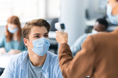 Temassız termometre tabancası olan öğretmen erkek öğrencinin ısısını ölçüyor.