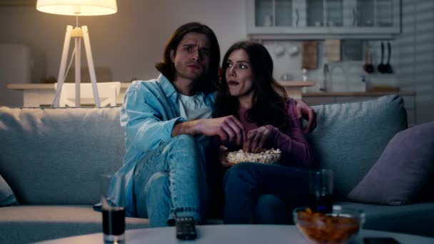 В помещении снимают молодую концентрированную пару, смотрящую фильм с попкорном, проводящую вечер дома, замедленную съемку — стоковое видео