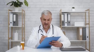 Doktor kameraya bakıp ofiste oturan notları alıyor.