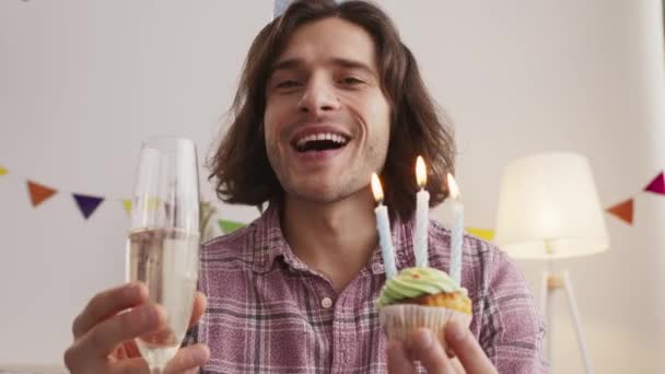 Portrait d'un gars heureux tenant un cupcake avec des bougies allumées et buvant du champagne dans un verre, souriant à la caméra pov — Video