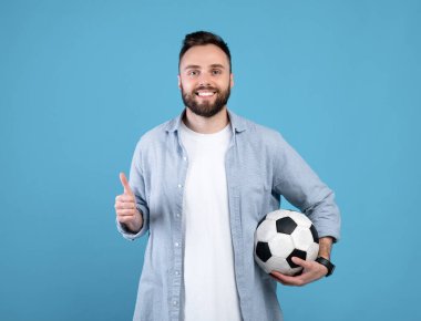 Futbol topu tutan neşeli genç adam, başparmak hareketi yapıyor, mavi stüdyoda spor öneriyor.