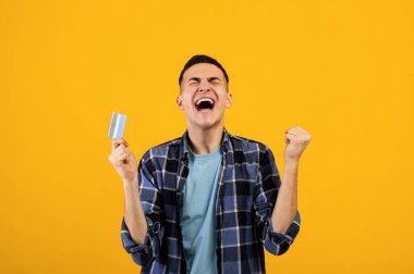 Kredi kartı ile heyecanlı genç adam çığlık atıyor ve turuncu stüdyo arka planında EVET hareketi yapıyor.