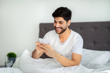 Mutlu Arap adam yatakta akıllı telefon kullanıyor, sabah uyanıyor ve internette geziniyor.