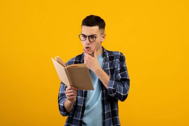 Gözlüklü, ders kitabı okuyan, turuncu stüdyo arka planında sınava çalışmakta zorlanan genç bir öğrenci şok oldu.