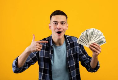 Heyecanlı milenyum adamı elindeki parayı işaret ederek turuncu stüdyo arka planındaki büyük zaferini gösteriyor.
