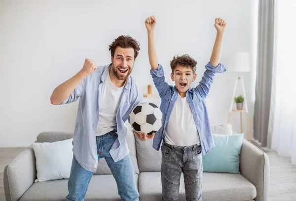 Emotional soccer fans. Cheerful dad and son cheering with football ball, watching match on tv