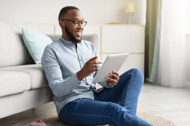 Mutlu siyah adam evde otururken dijital tablet kullanıyor.