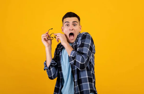 Horrified young guy yelling in panic, taking off his glasses over orange studio background