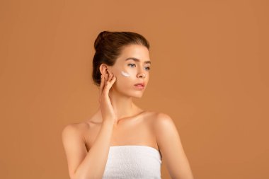 Kahverengi stüdyo arka planına yüz kremi süren şehvetli genç bir kadının portresi.