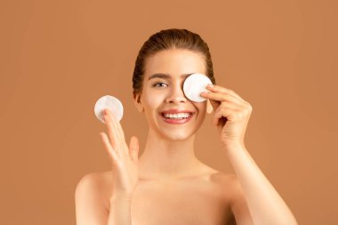 Yumuşak ciltli çekici bir kadın temizleyici tonik ya da mikiler suyu kullanıyor, kahverengi arka planda pamukla gözlerini kapatıyor.