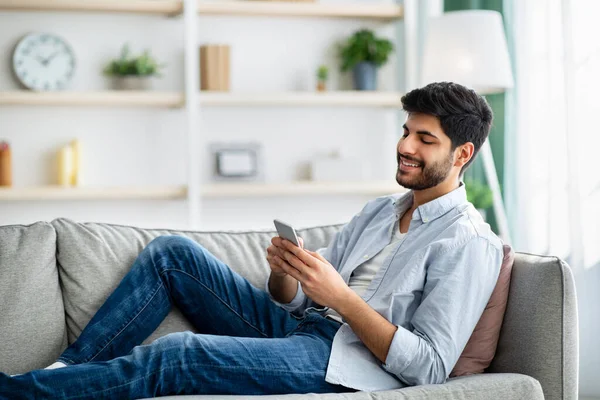 Mobile communication concept. Happy arab man using cellphone, browsing internet, sitting on couch at home, copy space