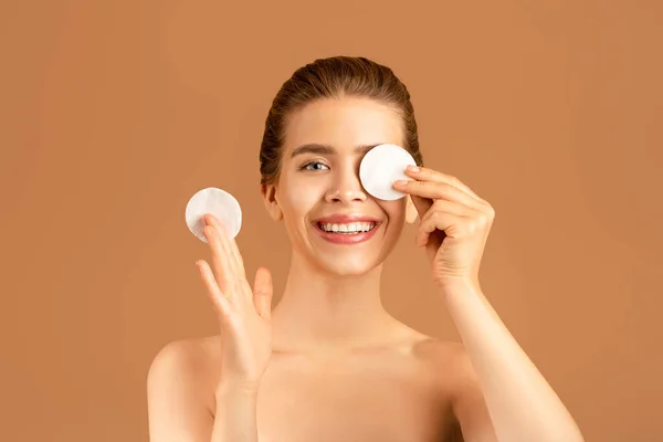 Charming woman with smooth skin using cleansing tonic or micellar water, closing eye with cotton pad on brown background