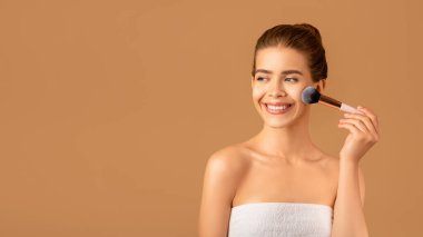 Genç cilt için organik makyaj. Şehvetli milenyum hanımefendisi allık sürüyor ya da kahverengi arka planda yüzüne pudra sürüyor.