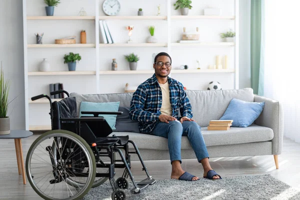 Happy disabled black guy sitting on sofa with books, empty wheelchair standing nearby, full length portrait