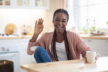 Neşeli siyah kadın mutfakta dinleniyor, kahve içiyor ve kameraya el sallıyor.