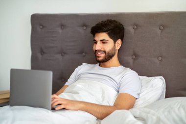 Uzaktan kumanda. Diz üstü bilgisayarda çalışan, evde yatakta otururken interneti karıştıran mutlu Arap serbest çalışanı..