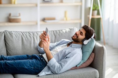 Boş zaman. Kablosuz kulaklıkla müzik dinleyen rahat bir Arap adam, rahat bir kanepede uzanıyor.