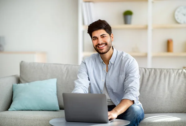Successful remote freelancer working distantly at laptop computer and smiling to camera, sitting on couch