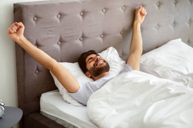 İyi uyuyan Arap adam uyanıyor ve ellerini uzatıyor, gözleri kapalı bir şekilde rahat bir yatakta yatıyor.